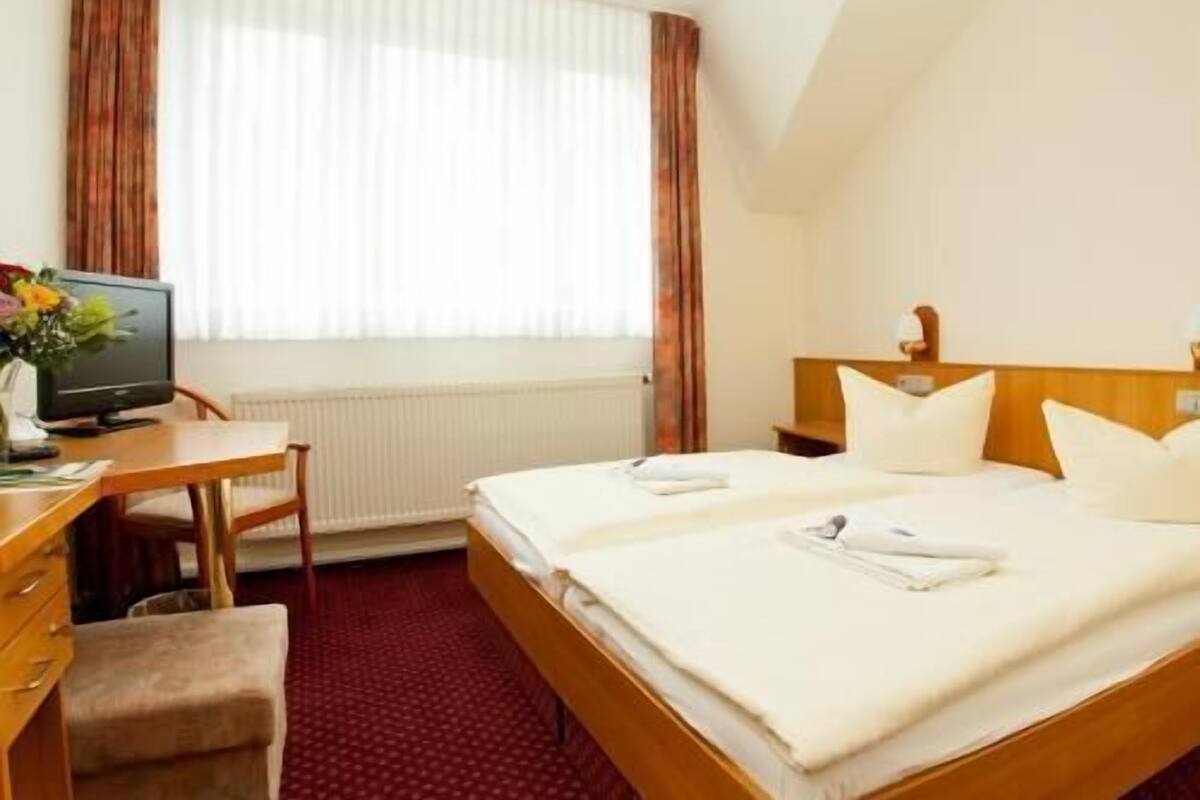 Hotellihuone, jossa on sänky, työpöytä tietokoneella, tuoli ja ikkuna, jossa on verhot.