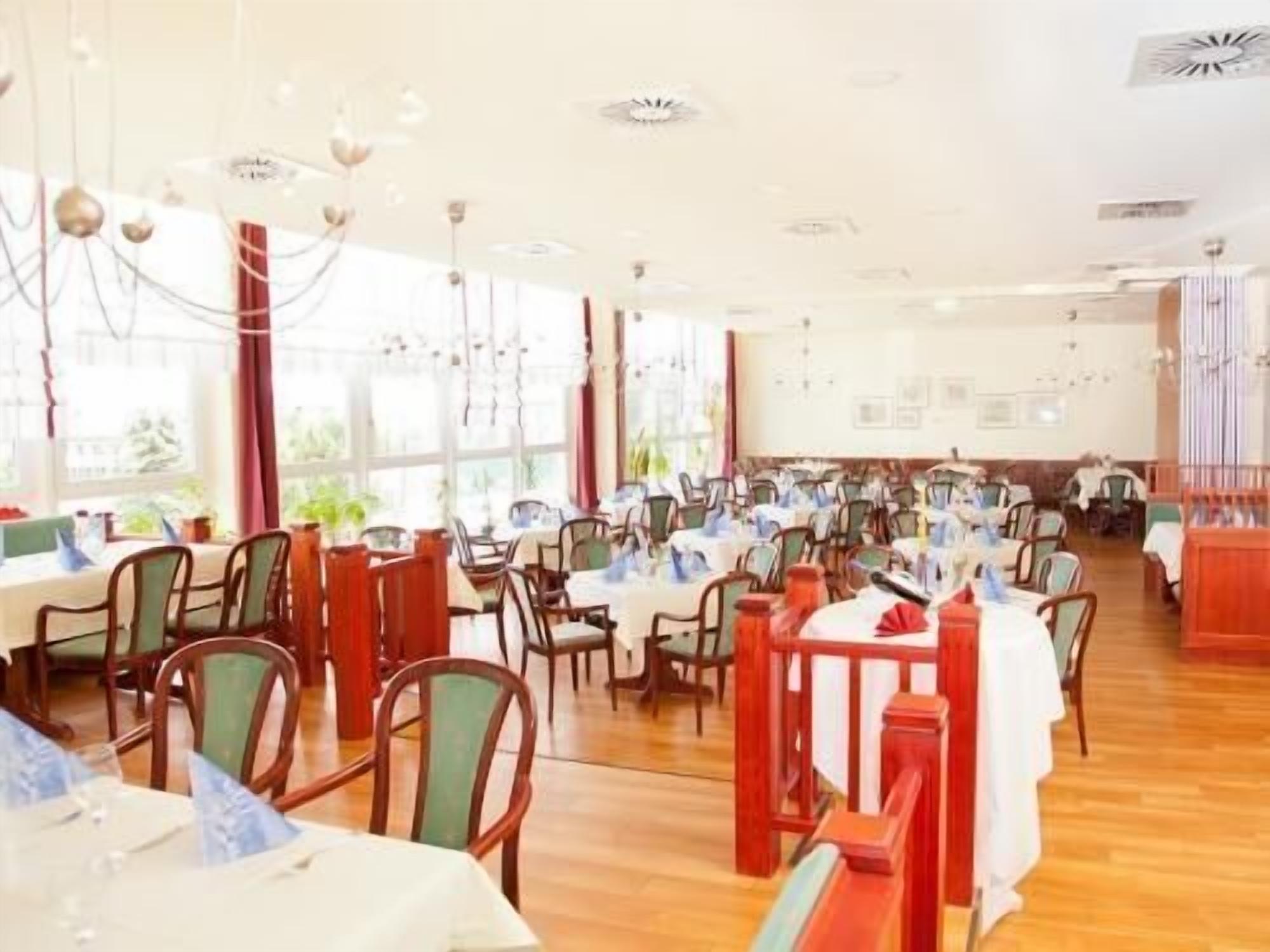 A spacious dining area with multiple tables and chairs, large windows allowing natural light, and a white ceiling with decorative elements.