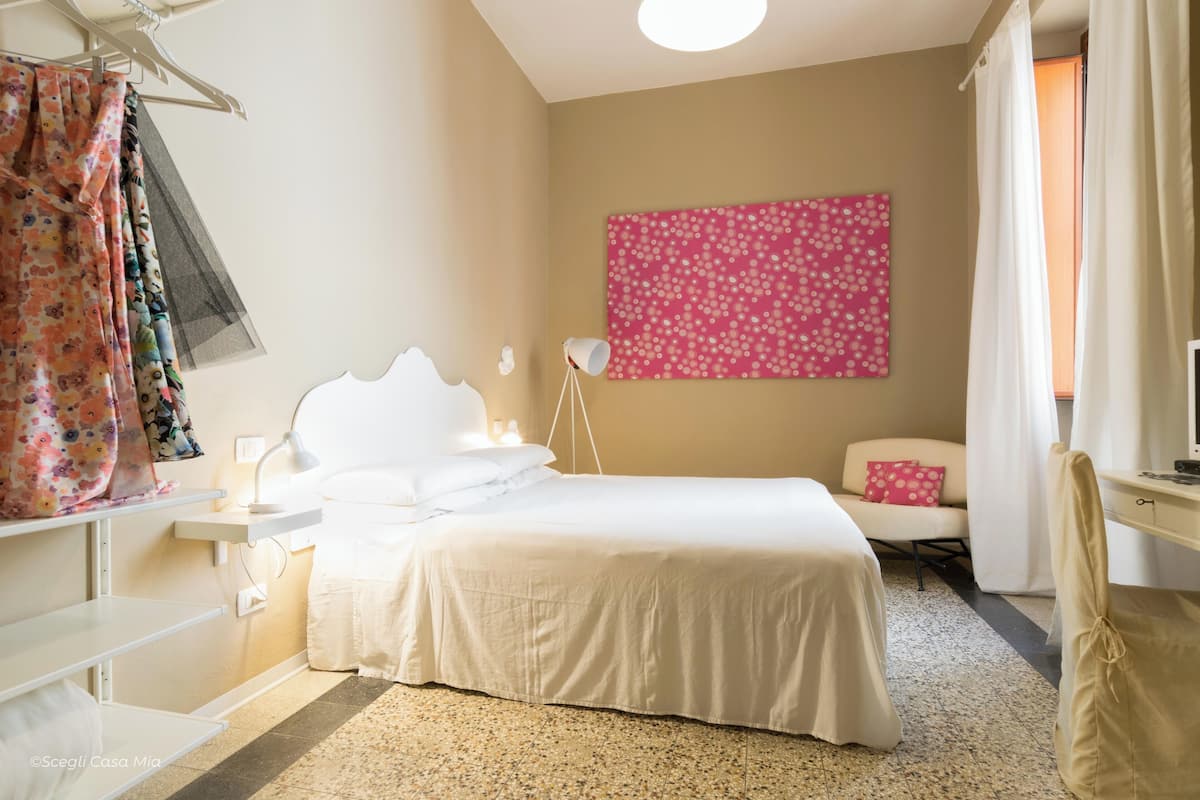 Een hotelkamer met een bed, nachtkastje, lamp, stoel en een roze wanddecoratie met patroon.