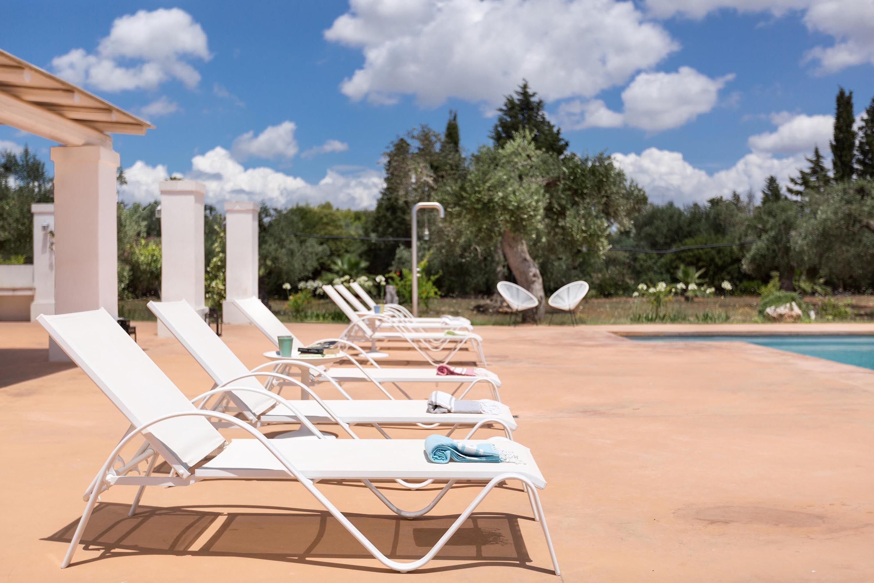 Fileira de espreguiçadeiras brancas com toalhas, piscina e uma pérgula num jardim.