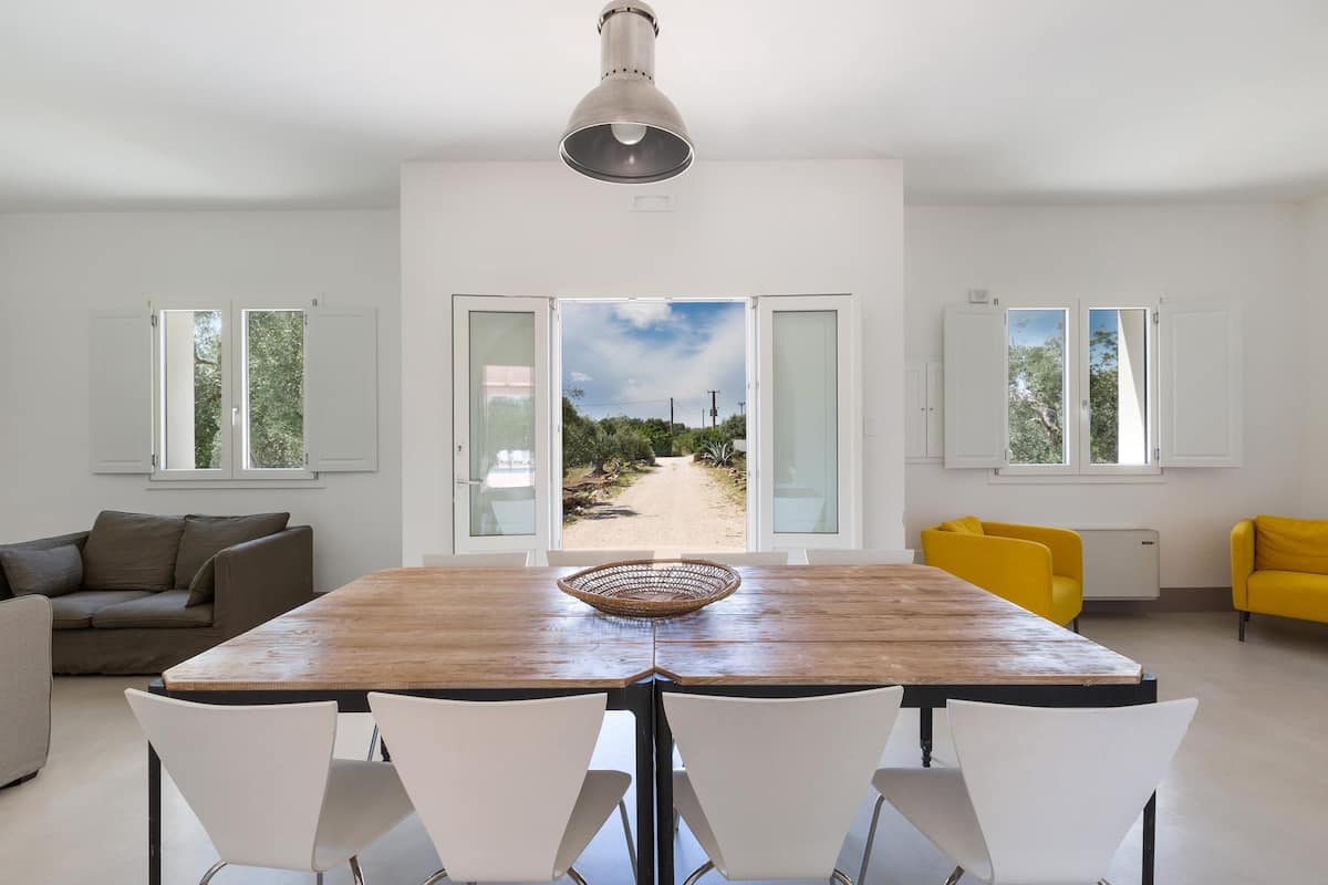 A dining area with a wooden table, white chairs, and a view through open doors to an outdoor path.