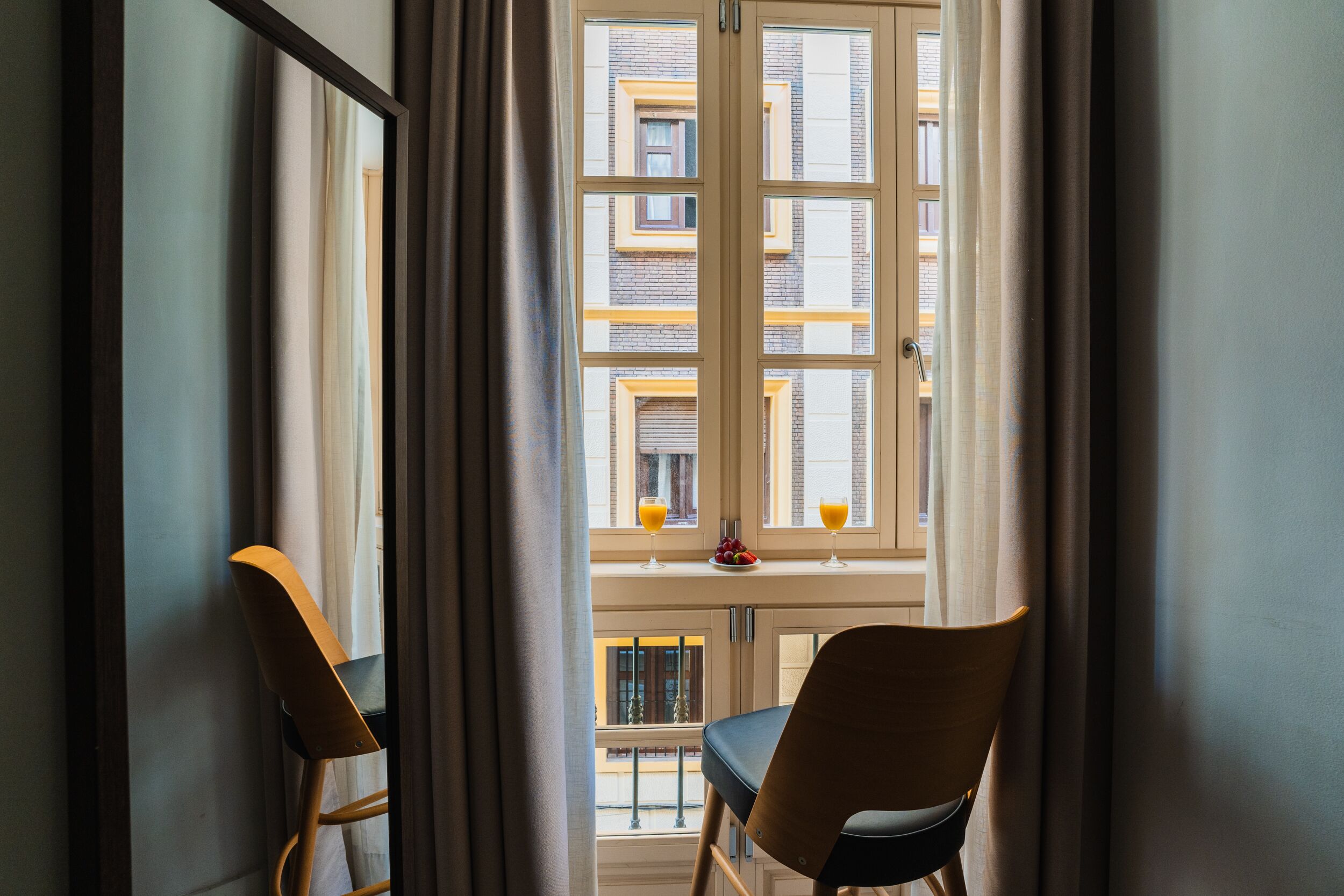 Une pièce avec vue sur un bâtiment, visible à travers une fenêtre ornée de rideaux. On y trouve deux chaises et une table sur laquelle sont disposés des fruits et des boissons.