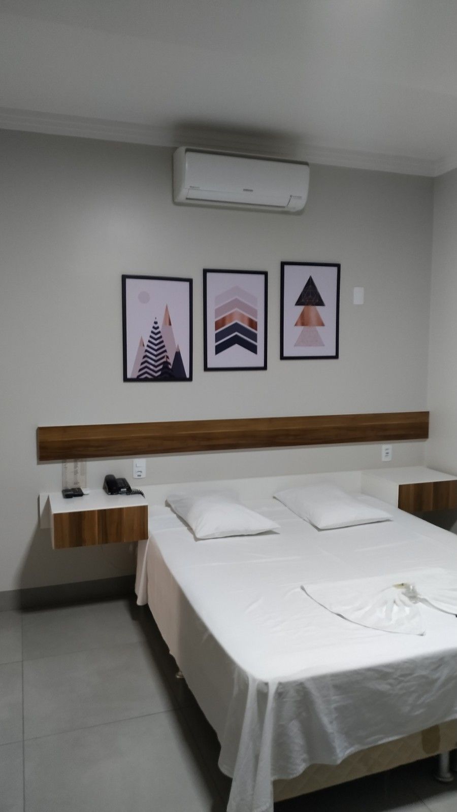 Een hotelkamer met een bed, twee nachtkastjes, een wandairconditioner en drie ingelijste kunstwerken.