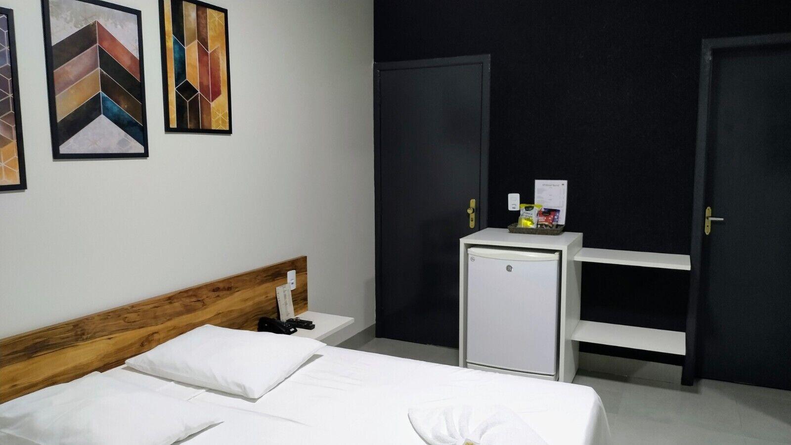 A hotel room with a wooden headboard bed, a small white bedside table, a mini fridge, and a black wardrobe.