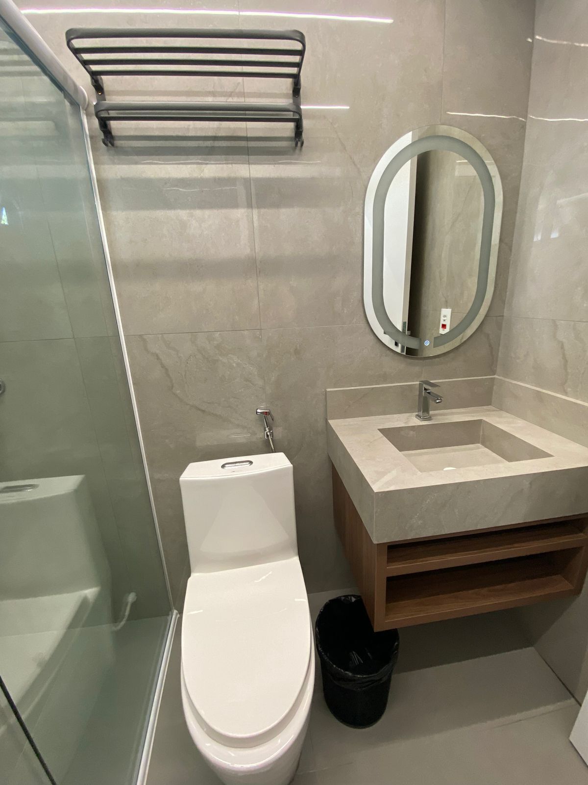 A modern bathroom with a glass shower enclosure, a white toilet, a sink with a mirror, and a black trash bin.