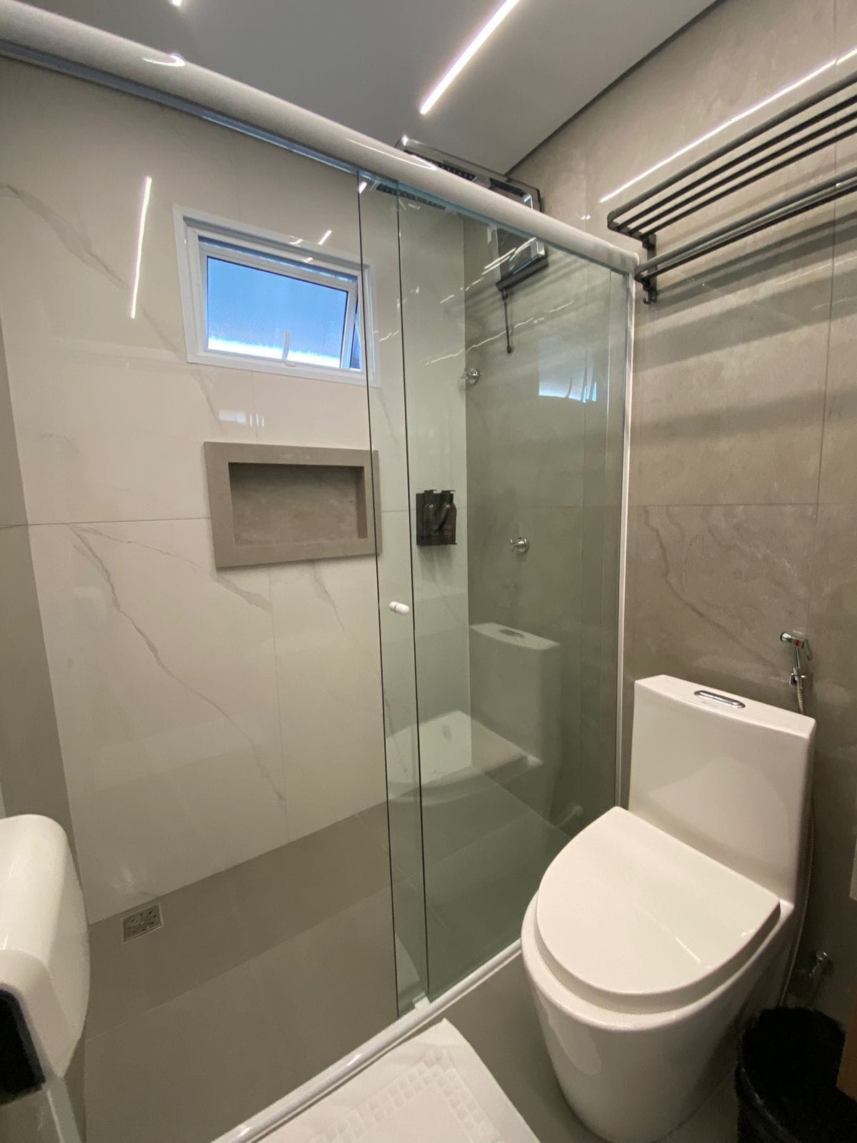 A modern bathroom with a glass-enclosed shower, a toilet, and a small window above the sink.