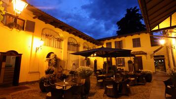 An outdoor patio with tables and chairs, a large umbrella, and a building with arches and a tiled roof.