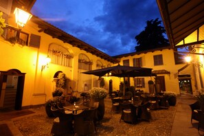 Un patio al aire libre con mesas y sillas, un paraguas grande y un edificio con arcos y techo de tejas.