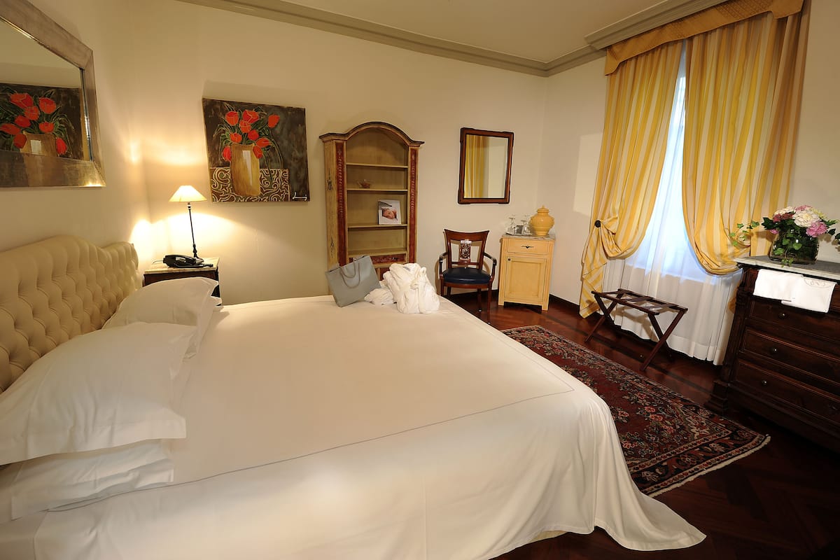 A hotel room with a large bed, a nightstand with a lamp, a wooden cabinet, a chair, and a desk with a vase of flowers.