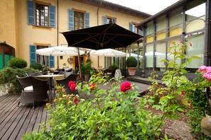 Un patio al aire libre con mobiliario de mimbre y sombrillas blancas, rodeado de macetas con plantas y flores.