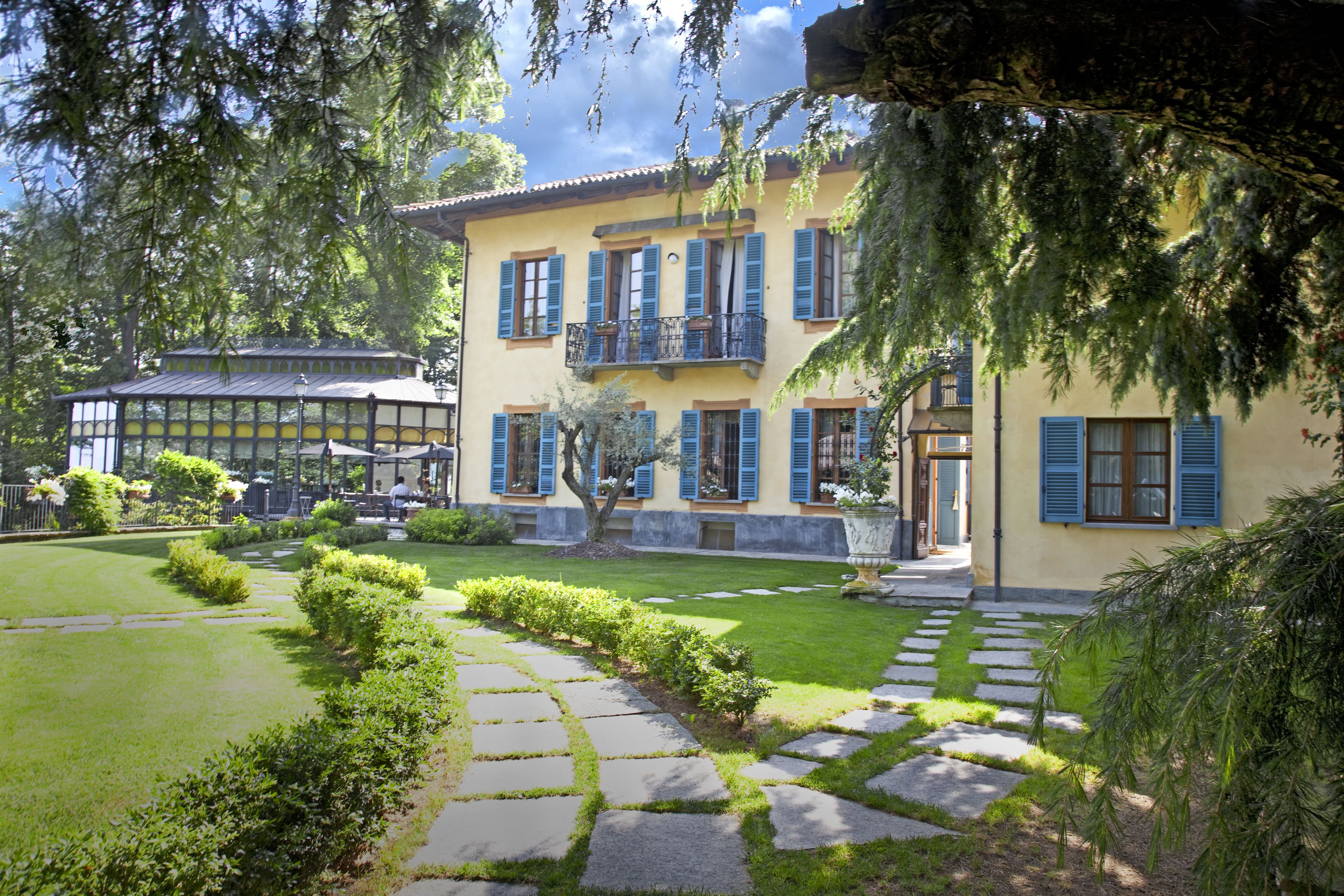 A two-story building with a yellow facade, blue shutters, and a balcony. There is a well-maintained lawn with a stone pathway leading to the entrance.