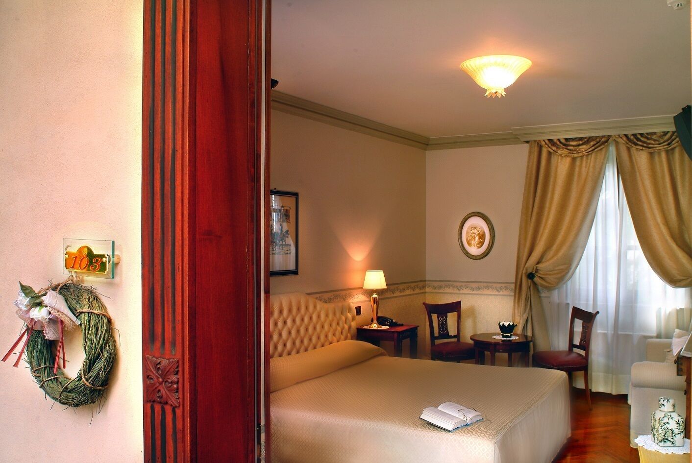 Een hotelkamer met een groot bed, een nachtkastje met een lamp, een stoel, een tafeltje met een decoratief object, en een raam met gordijnen.