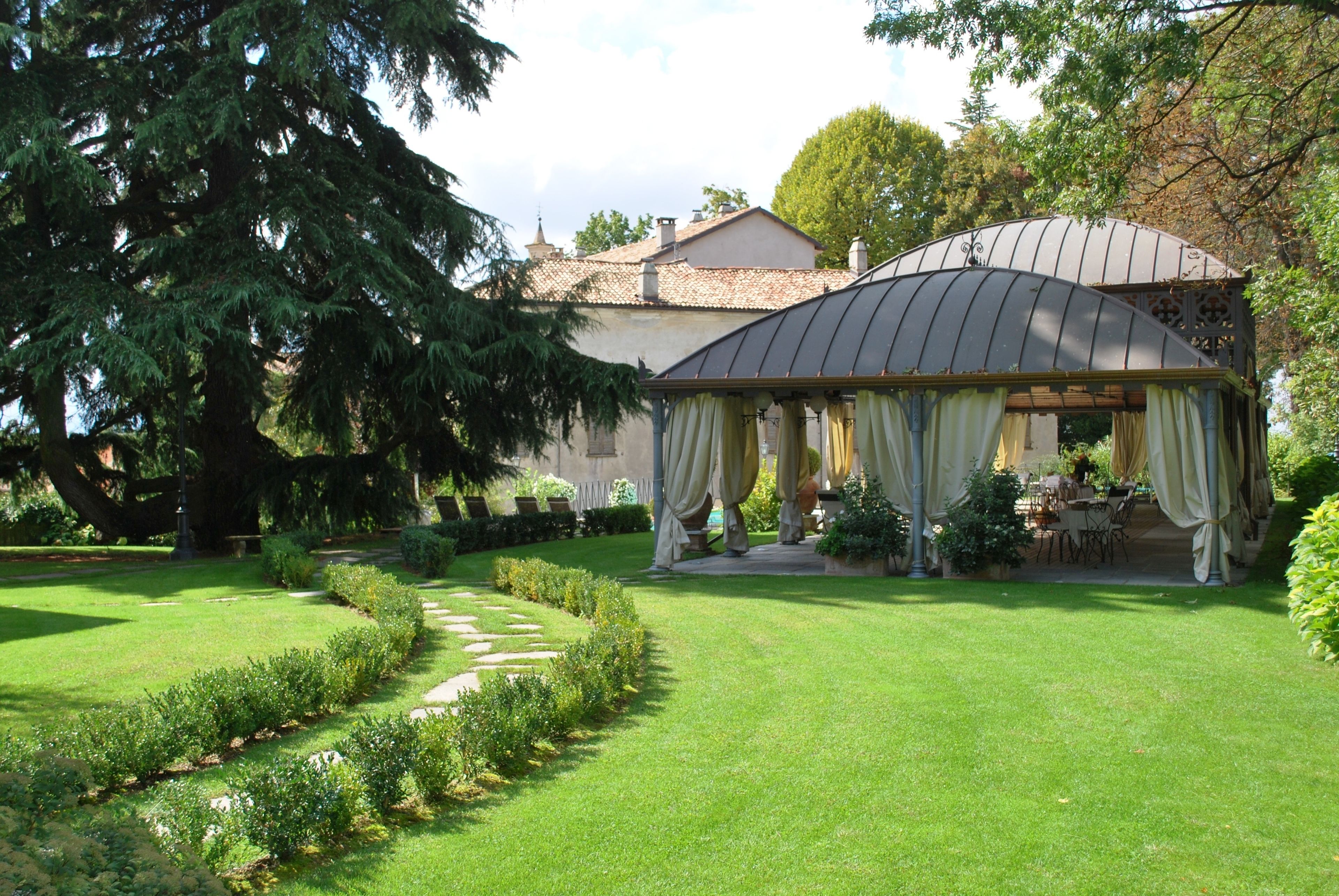 Um gazebo com telhado curvo e cortinas esvoaçantes, cercado por um gramado bem cuidado e um caminho de pedra.