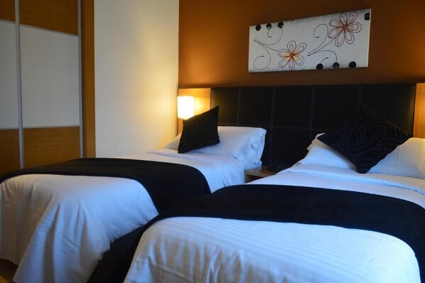 Habitación de hotel con dos camas, cada una con ropa blanca y almohadillas negras. Hay una obra de arte colgada en la pared sobre las camas, y una mesita de noche con una lámpara.