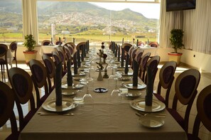 Una larga mesa de comedor dispuesta para una comida, con sillas a ambos lados, plantas en macetas y vistas a un pueblo y montañas.