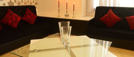 A modern living room with a glass coffee table, black and red patterned sofa, and three lit candles on a shelf.