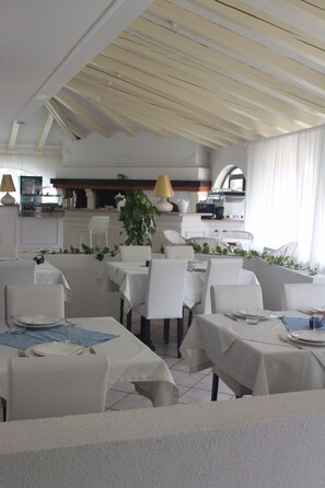 A dining area with white tables and chairs, a kitchen in the background, and a plant in the center. - Hotel La Settima Luna (Umag)