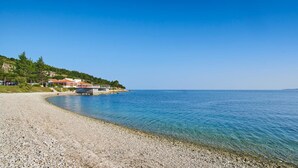 A pebble beach with clear water, a building with red roofs, and a hilly backdrop. - Hotel La Settima Luna (Umag)