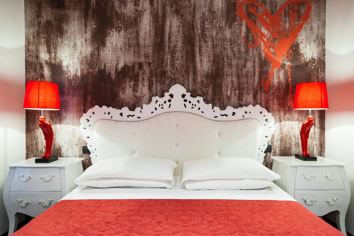 Una cama con cabecera blanca, dos mesitas de noche con lámparas rojas y una pared texturizada con diseño de corazón.