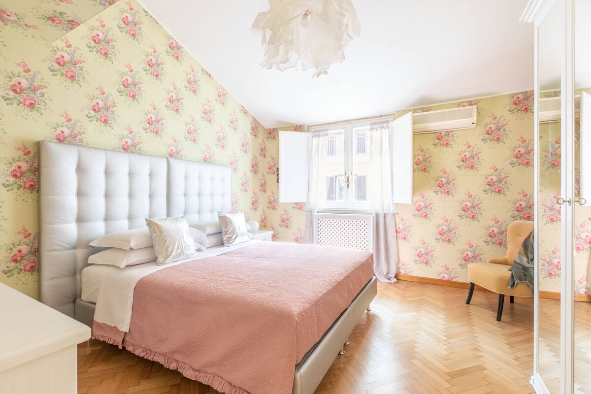 Un dormitorio con cabecera capitonada, cama con colcha rosa, papel tapiz floral y suelo de madera.