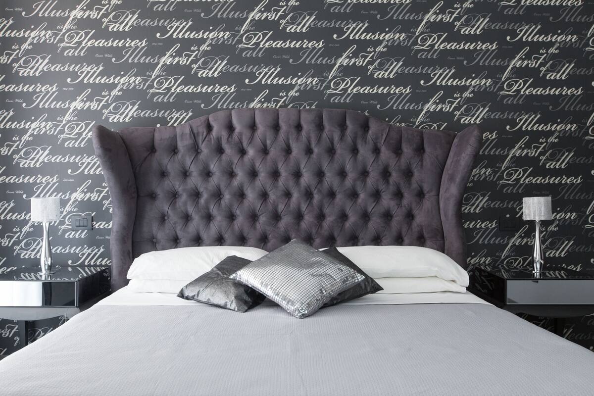 Un dormitorio con cabecera capitonada, dos almohadas, una mesita de noche con lámpara y una pared con texto repetido.