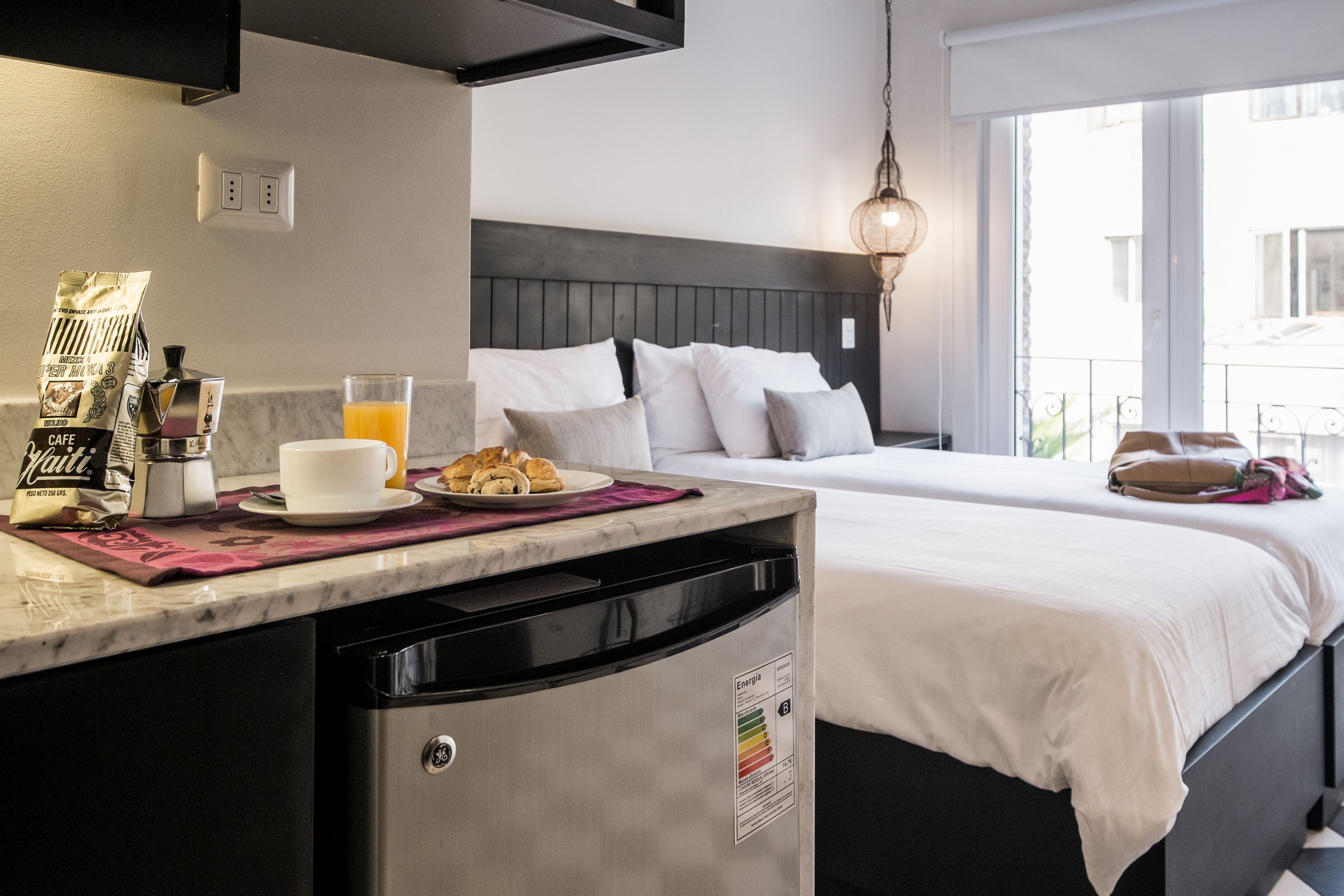 A hotel room with a marble countertop, a bed with white bedding, a bedside table with a lamp, and a view of the outside through a large window.