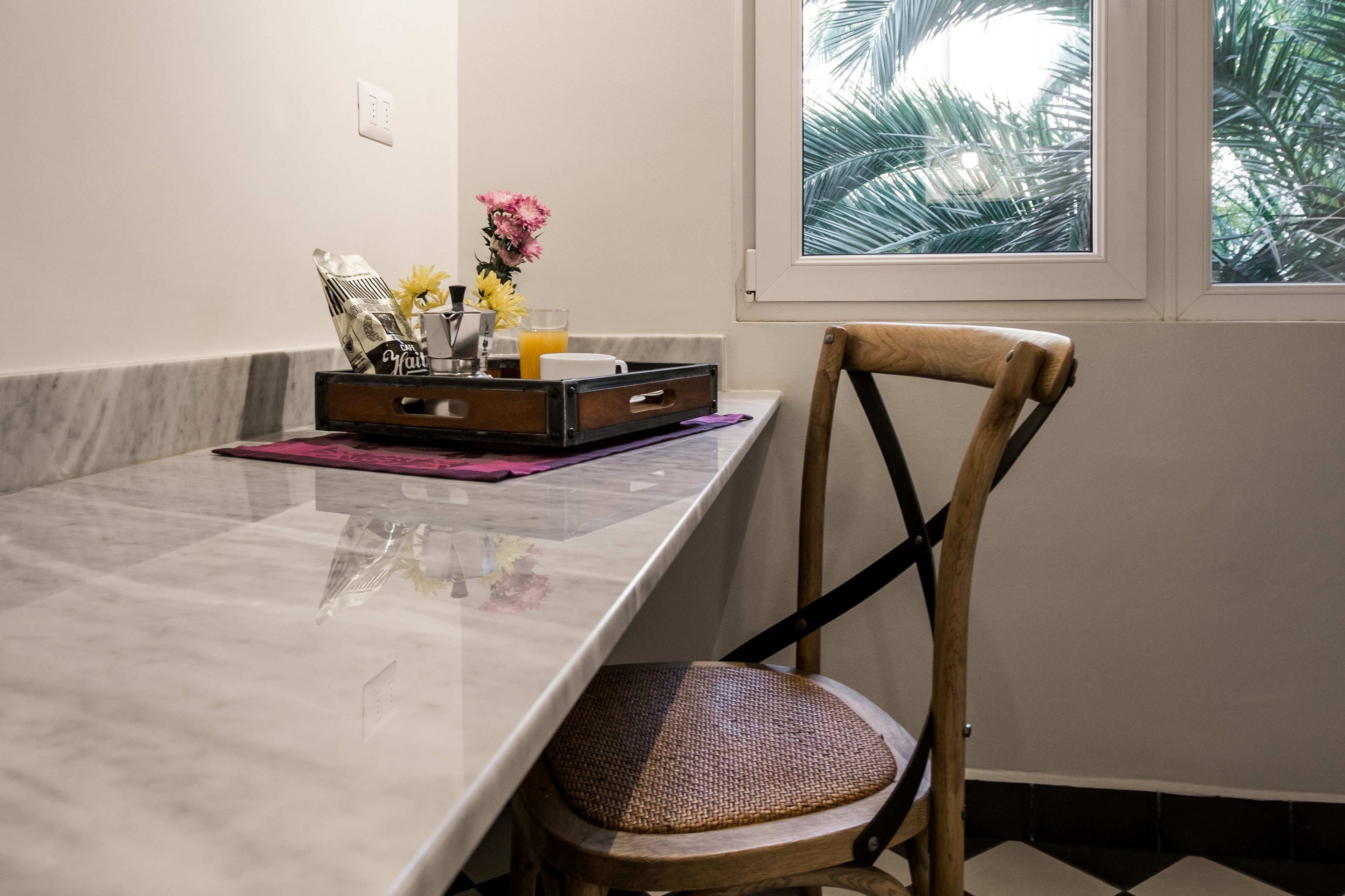 Un coin repas avec une table en marbre, une chaise en bois, un plateau contenant des collations et une boisson, et une fenêtre donnant sur des palmiers.