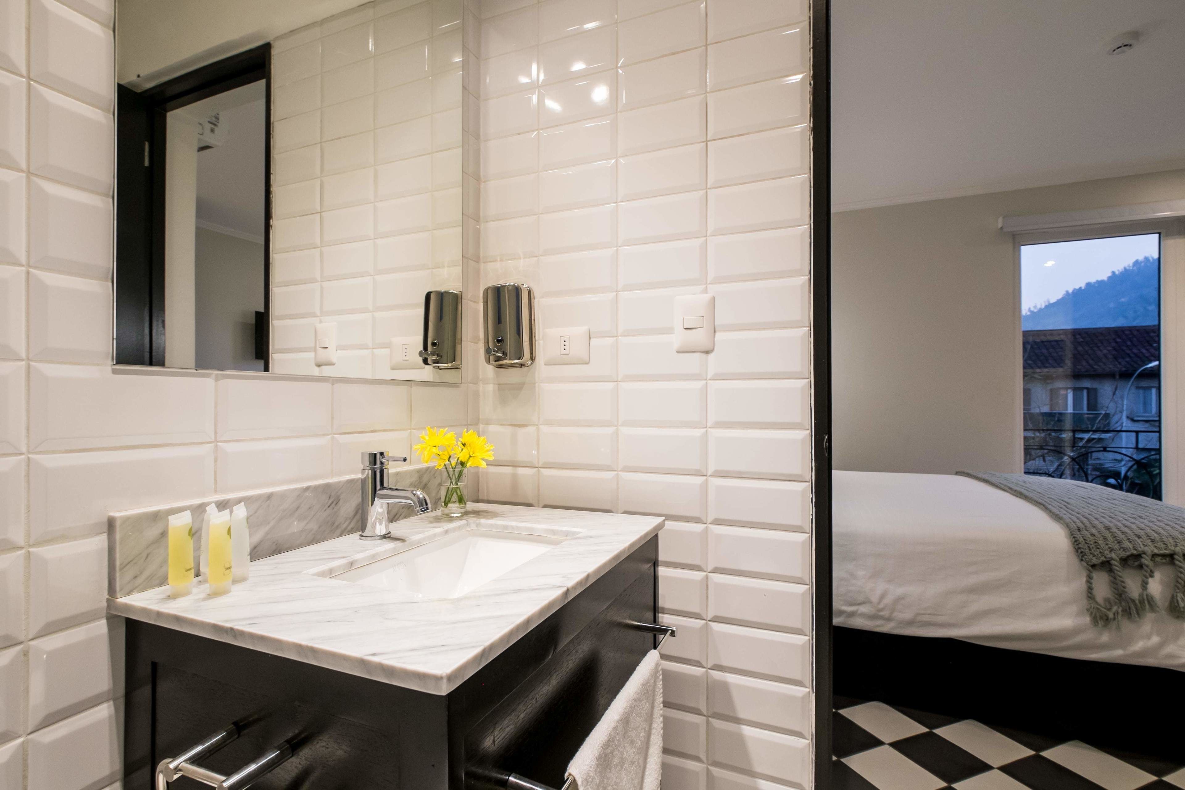 A modern bathroom with a large mirror, white tiled walls, a marble sink, and a small vase with yellow flowers.