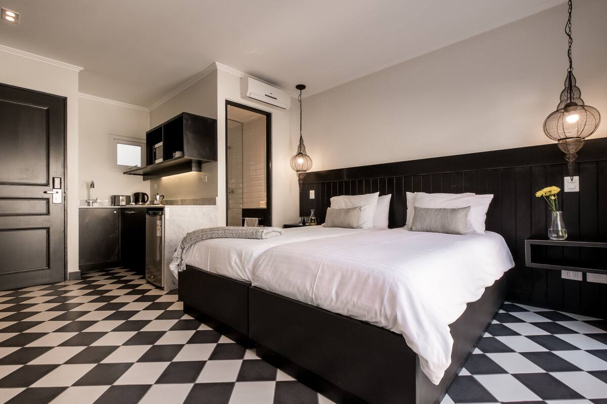 A modern bedroom with a large bed, a small bedside table, a nightstand, a hanging light fixture, and a bathroom visible through an open door.