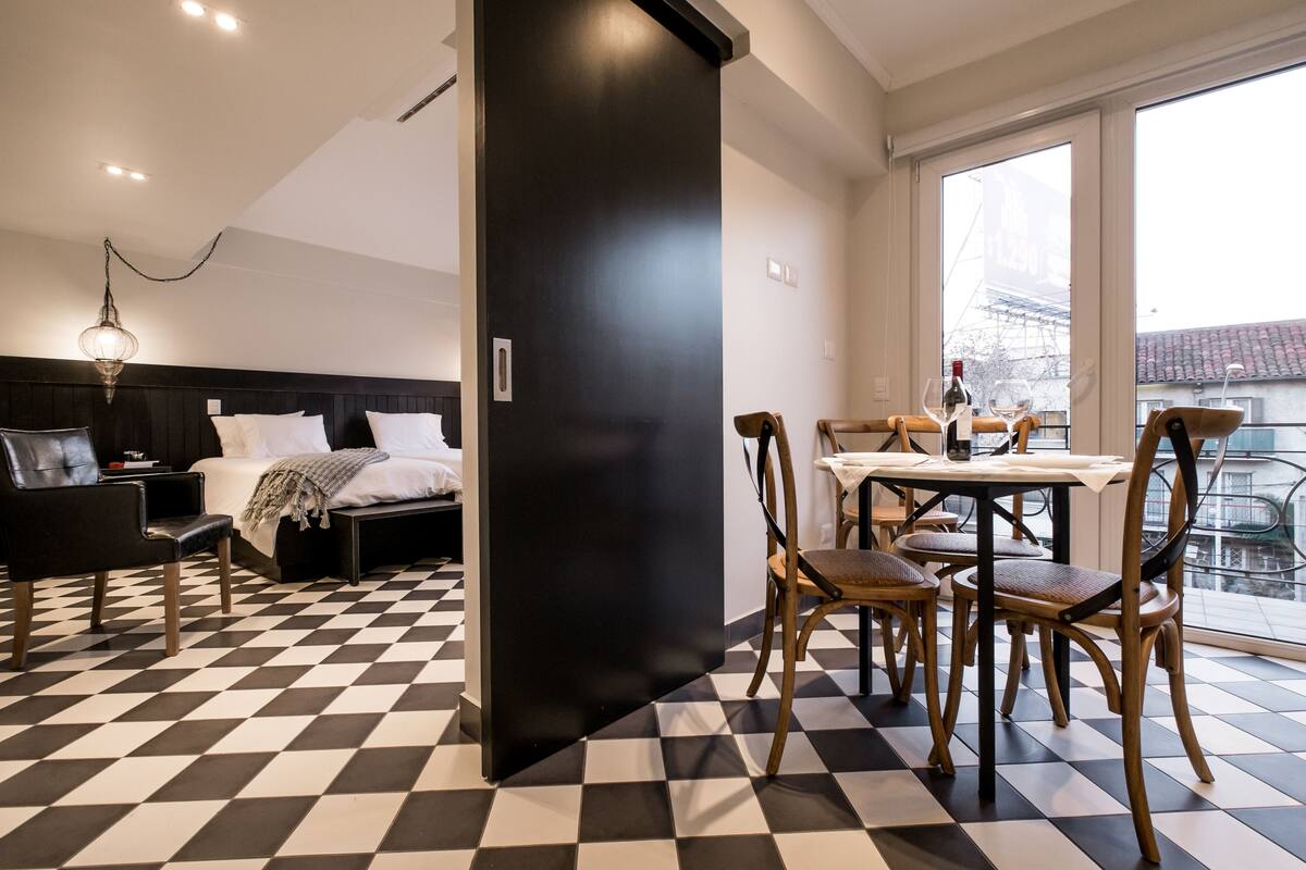 A hotel room with a checkered floor, a bed with white linens, a black headboard, a dining table with chairs, and a view of the city through large windows.