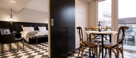 A hotel room with a checkered floor, a bed with white linens, a black headboard, a dining table with chairs, and a view of the city through large windows.