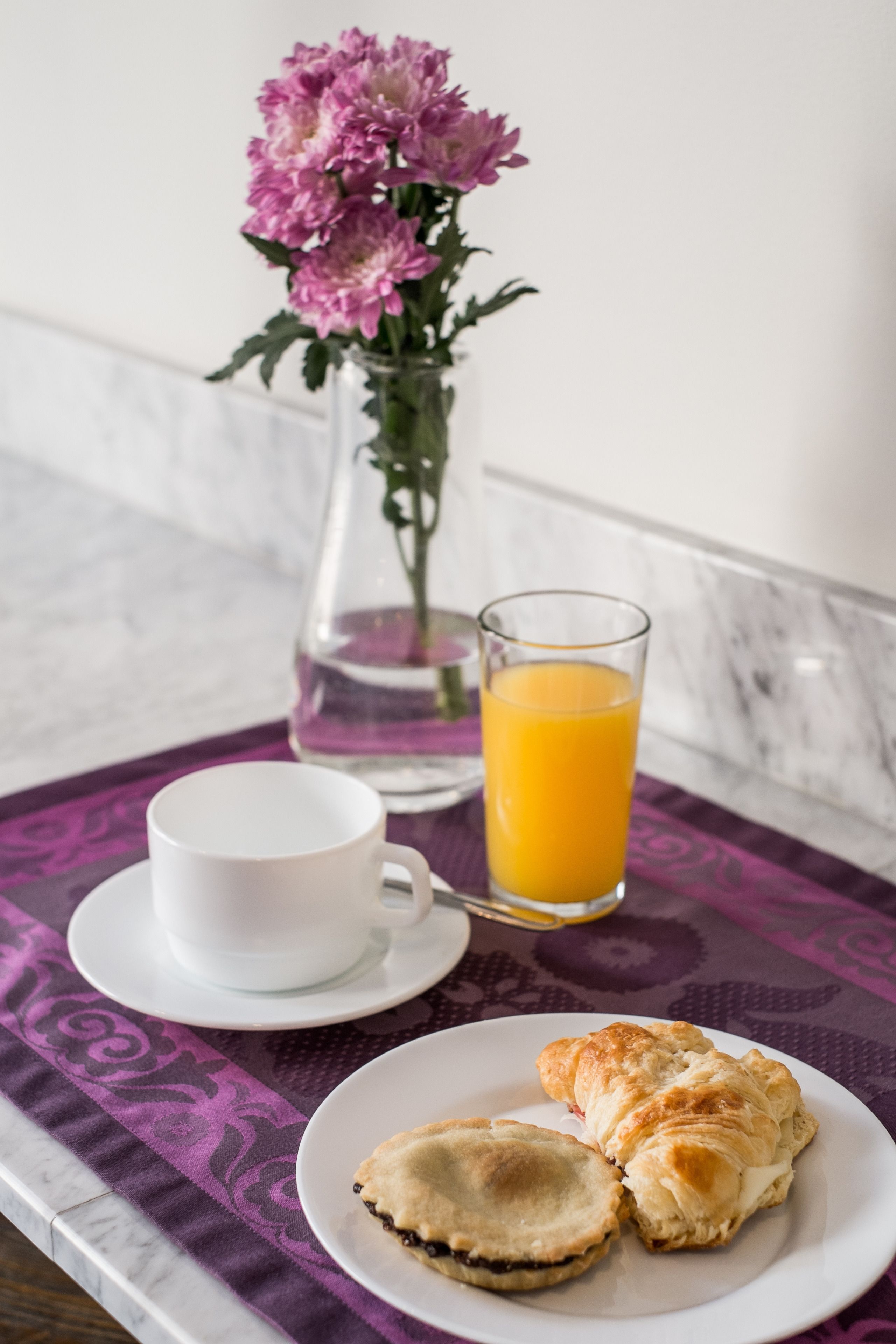 A breakfast tray with a cup, a glass of orange juice, and two pastries on a table with a purple tablecloth.