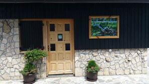 Eine Holztür mit Glasfenster, eingerahmt von zwei Kübelpflanzen mit roten Blüten, vor einer Steinwand mit einer schwarzen Markise.