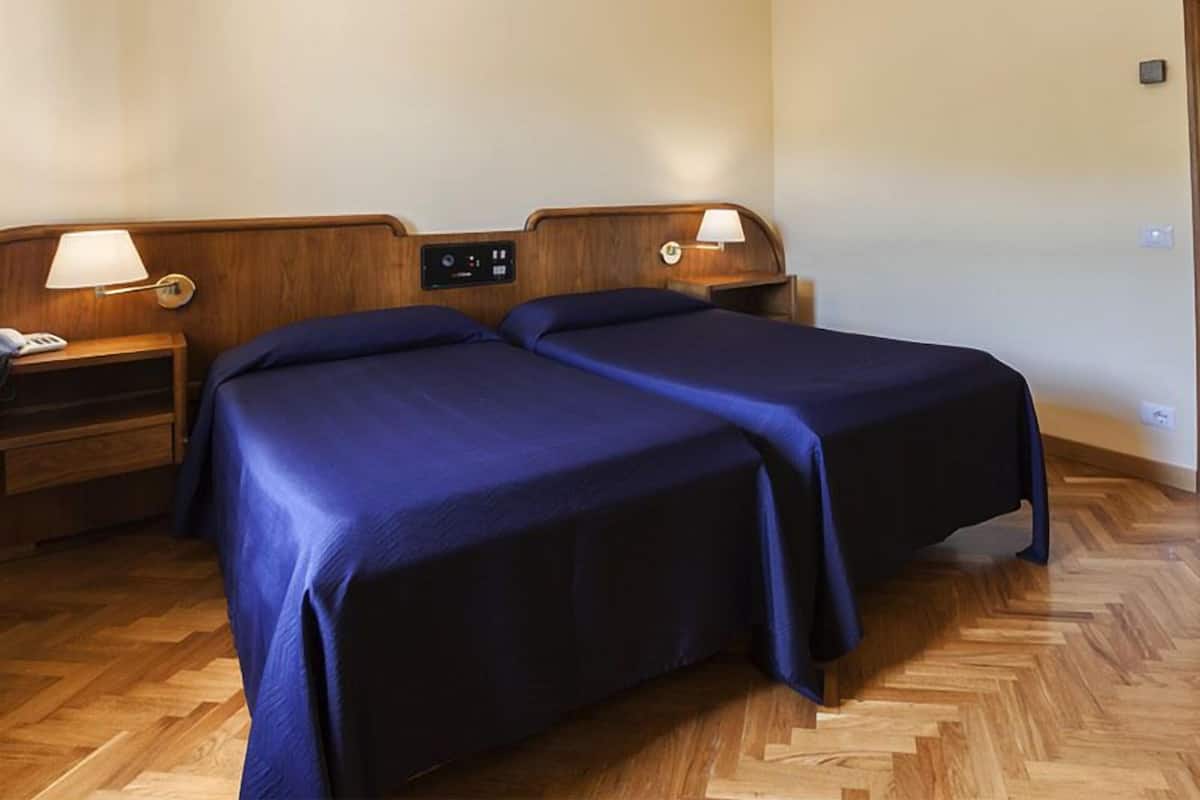 A hotel room with two beds, wooden headboards, bedside tables with lamps, and a control panel on the headboard.