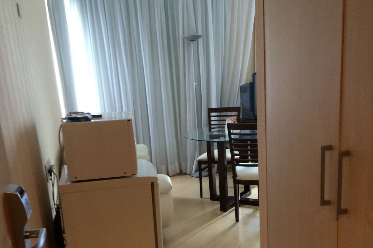 A hotel room with a wooden floor, a bed, a desk with a chair, a television, and a window with curtains.