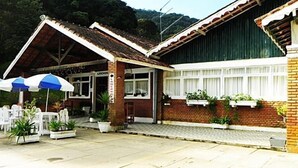 A building with a brick facade, a covered porch with white chairs, and potted plants. - Hotel H Santo Antonio do Pinhal (Monteiro Lobato)
