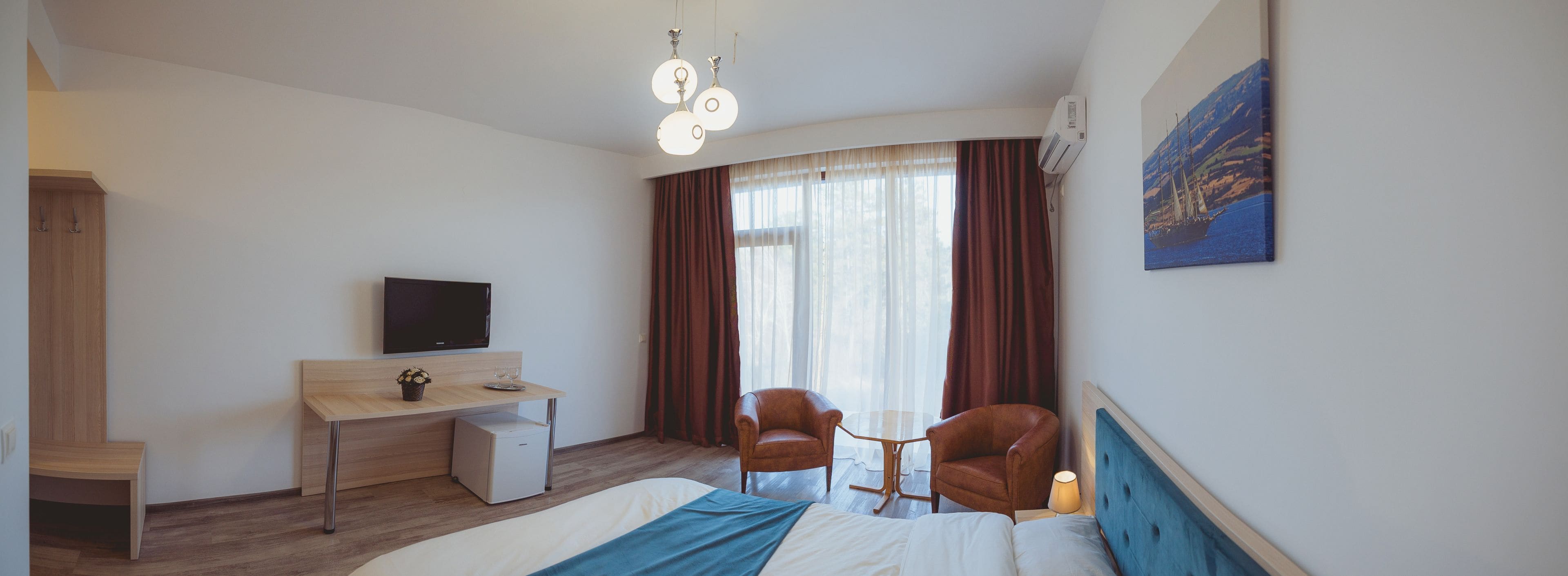 A hotel room with a bed, a desk with a TV, a chair, a small table, and a window with curtains.