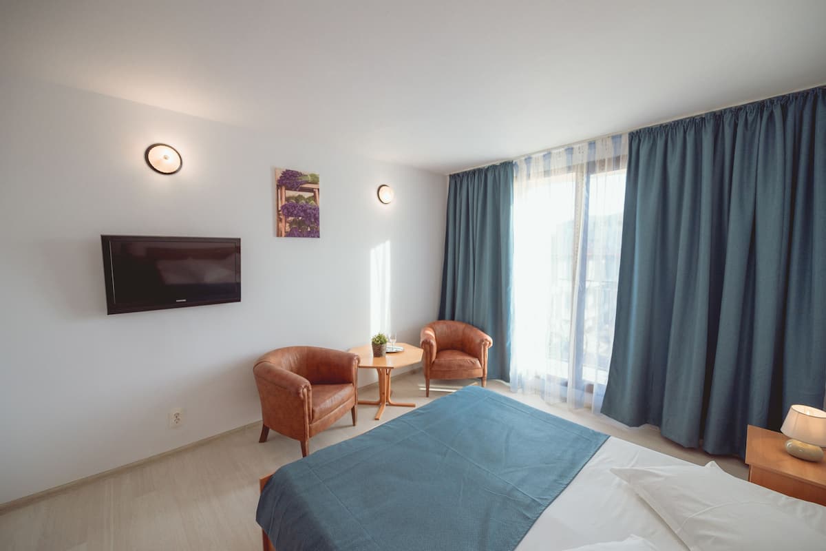 A hotel room with a bed, two armchairs, a small table, a TV mounted on the wall, and a window with curtains.