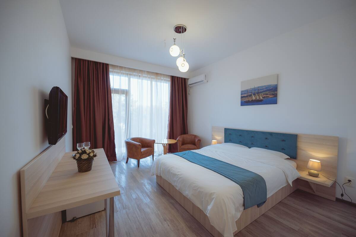 A modern hotel room with a large bed, a desk with a lamp, a chair, and a painting on the wall.