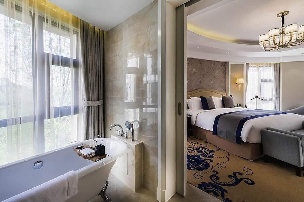A modern hotel room with a bathtub, a large bed, a chandelier, and a view of trees through the window.