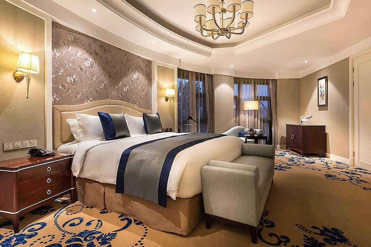 A spacious hotel room with a large bed, a chandelier, a desk, and a view of the outdoors.