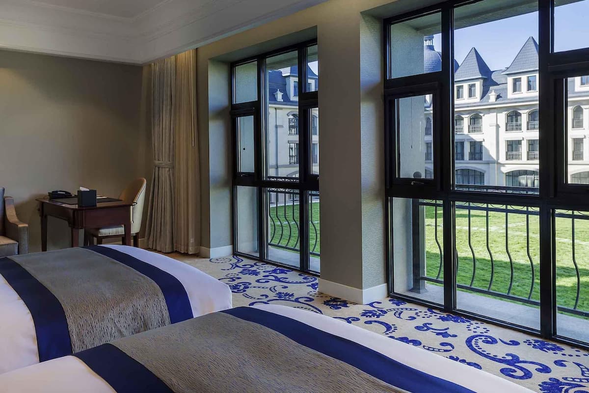 A hotel room with two beds, a desk, and large windows offering a view of a building and a lawn.