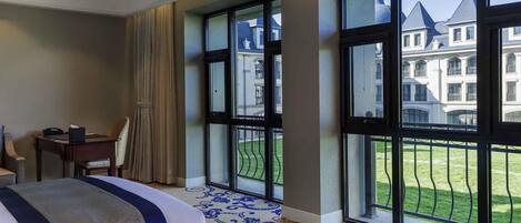 A hotel room with two beds, a desk, and large windows offering a view of a building and a lawn.