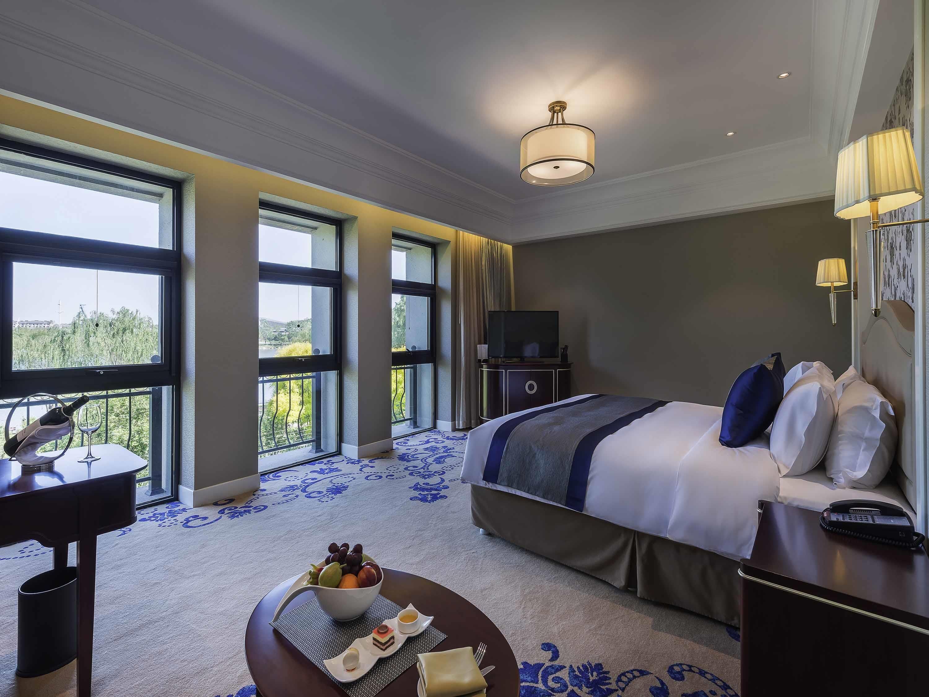 A hotel room with a large bed, a desk, a television, and a view of greenery through large windows.