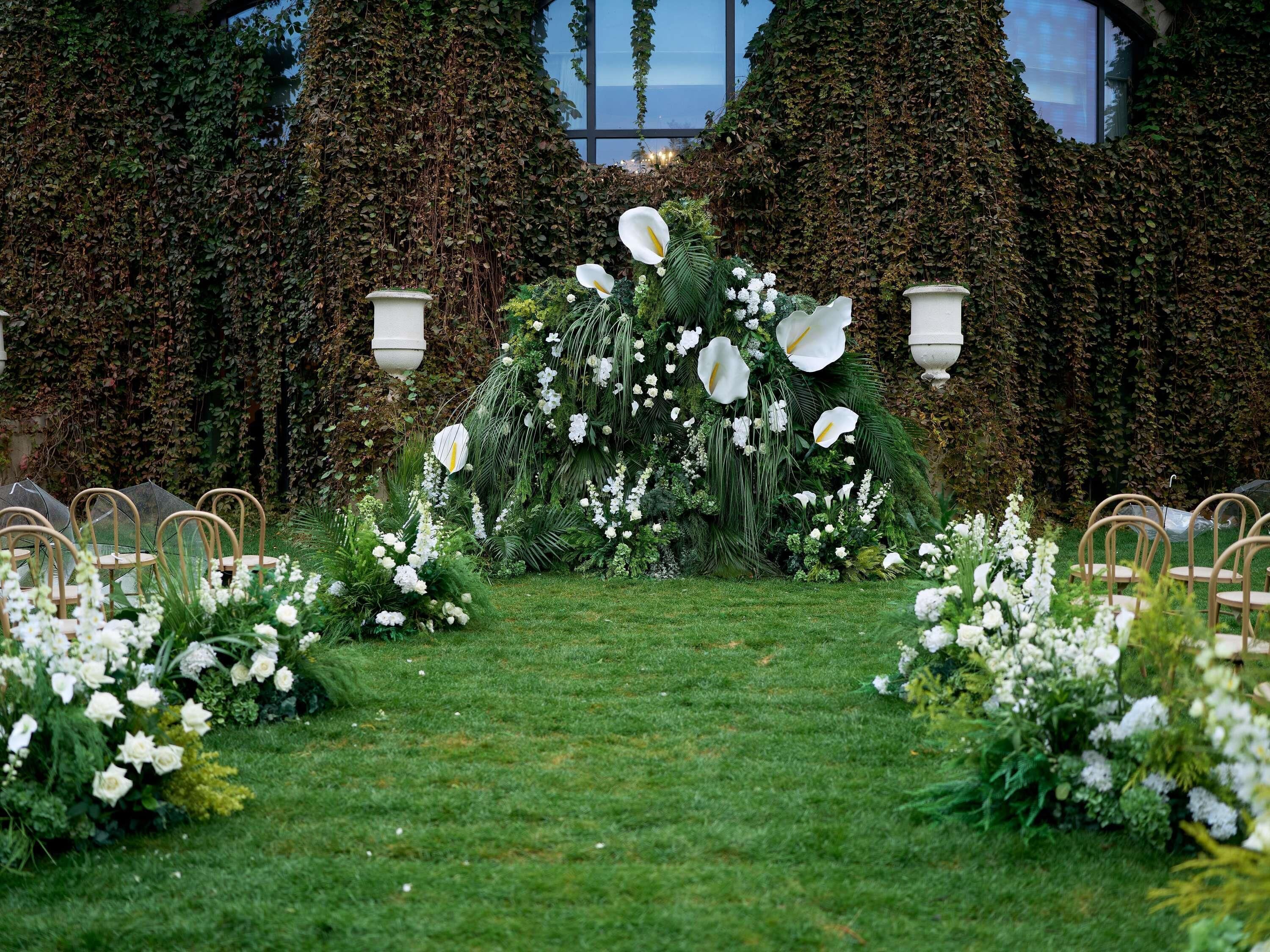 An outdoor wedding arch with white flowers and greenery, surrounded by chairs.