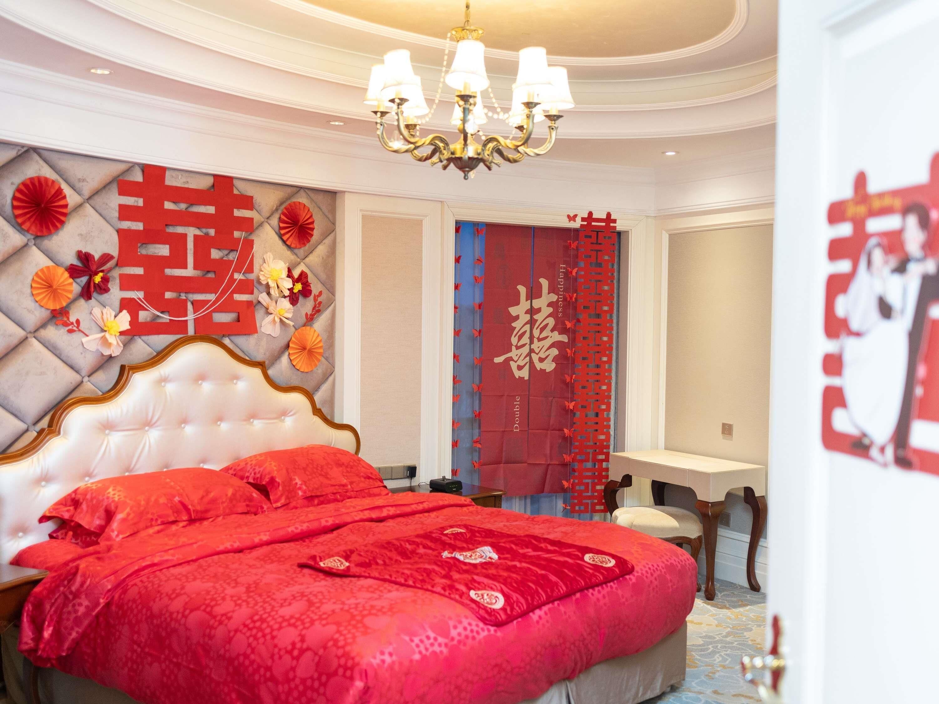 Un dormitorio con una cama grande, una araña y elementos decorativos, incluyendo una pared roja con caracteres chinos y una cortina roja con caracteres chinos.