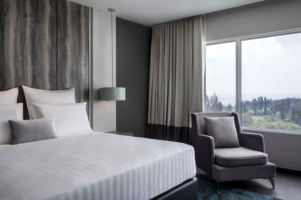 A modern hotel room with a large bed, a chair, a lamp, and a view of the outdoors.