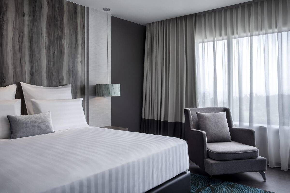 A modern hotel room with a large bed, a chair, a wall-mounted lamp, and a view through sheer curtains.