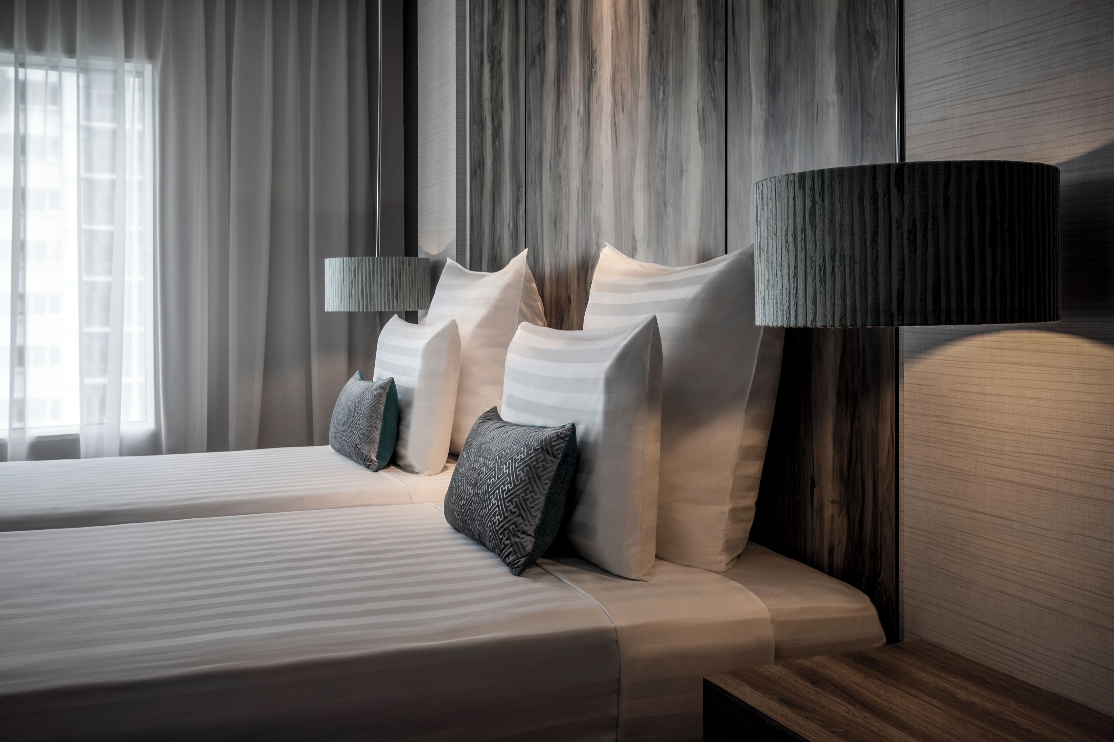 A double bed with a wooden headboard, two bedside lamps, and a view of a cityscape through a window with sheer curtains.