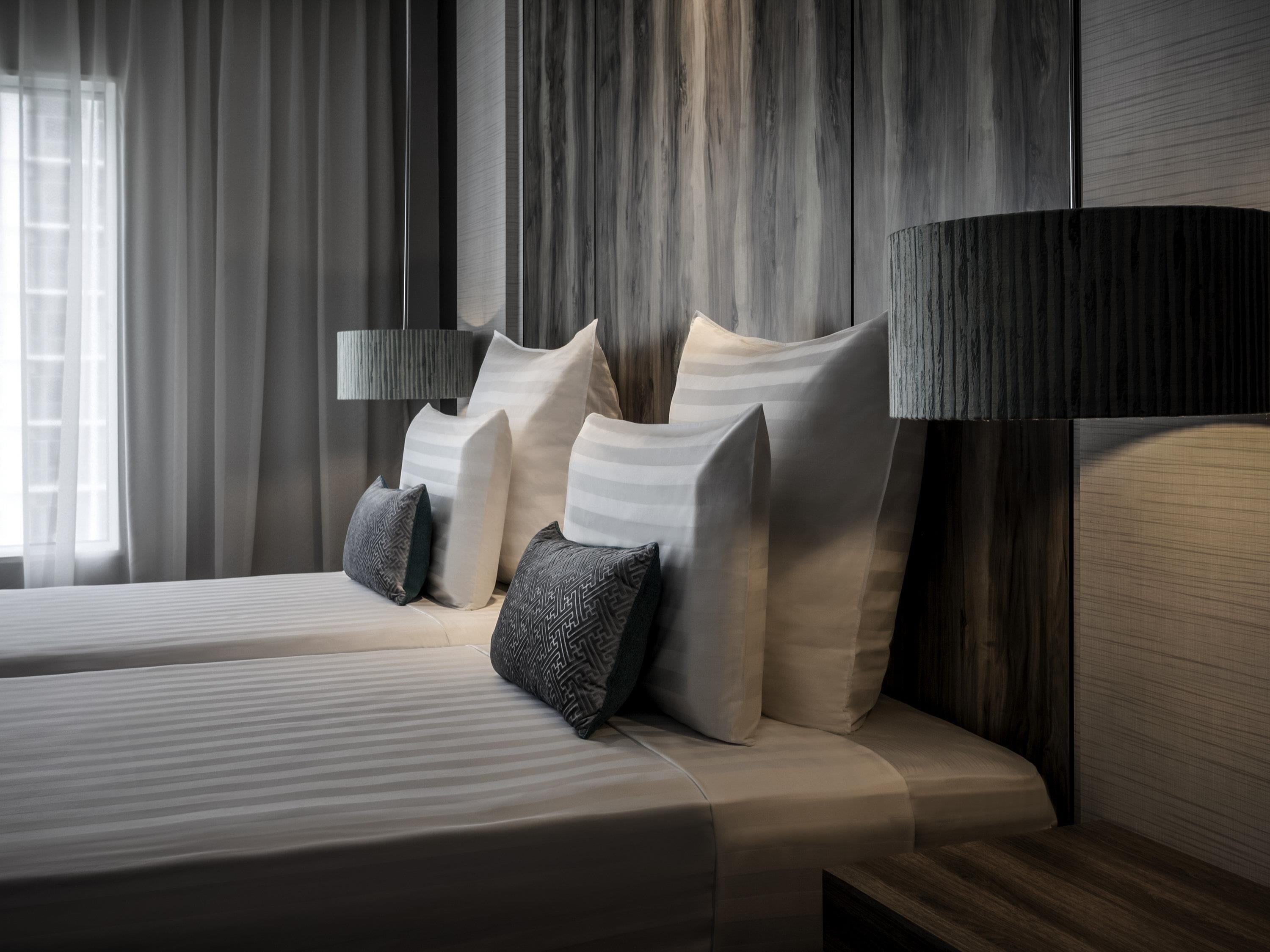 Moderni hotellihuone, jossa on suuri sänky, useita tyynyjä, yöpöytälamppu ja ikkuna, jossa on verhot.